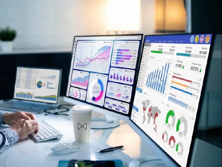 Two monitors of dashboards on a desk beside a Business X cup