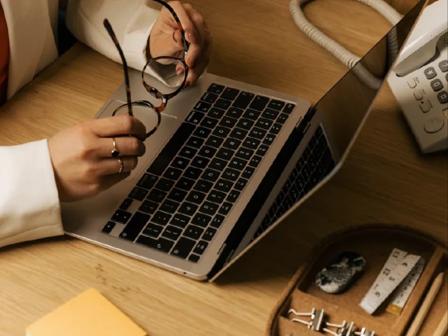 A woman holding her glasses while working at a laptop on a wooden desk