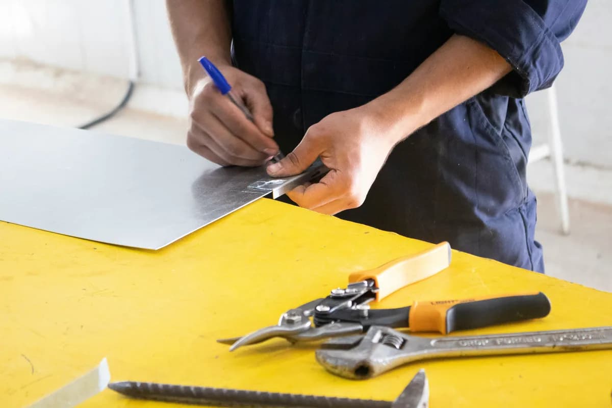 A metalworker marking a steel sheet beside pliers and a wrench on a yellow bench