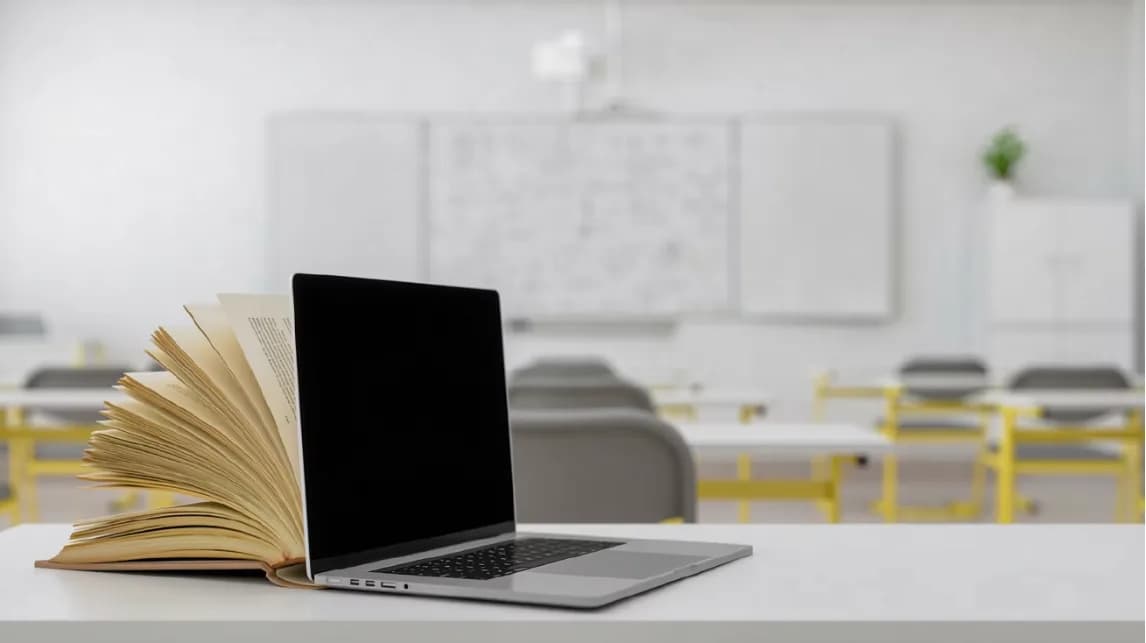 An open book and a laptop on a desk in an empty classroom