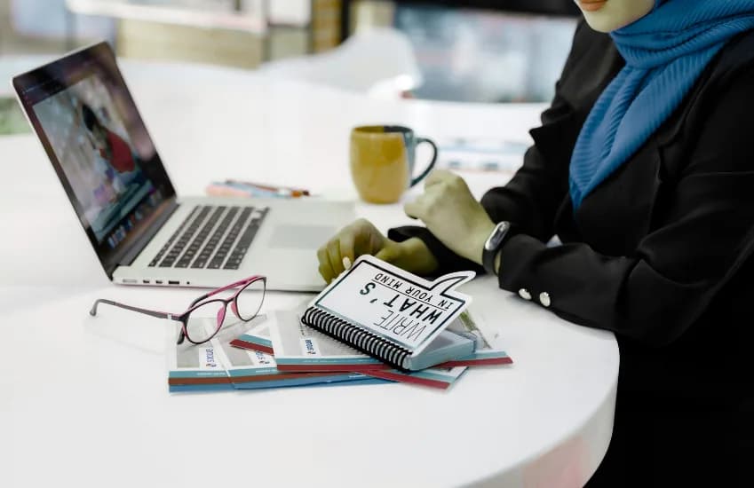 A woman in a headscarf at a laptop with notebooks and a coffee cup